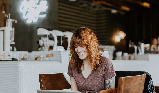 A woman with shoulder-length red hair and a casual T-shirt works on her laptop at a wooden table inside a modern café.