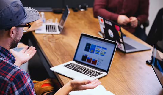 A man in a plaid shirt and cap gestures during a team meeting, with laptops open on the table showing colorful design mockups.