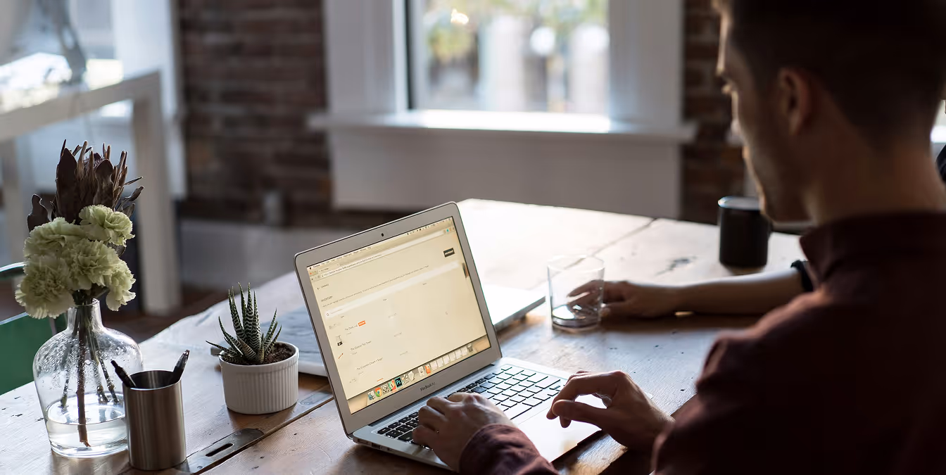 A man sits at a wooden table in a sunlit office, working on a MacBook with a minimalist website dashboard open.
