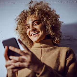 A smiling young woman with curly blonde hair wearing a tan turtleneck, leaning against a wall and looking at her smartphone.