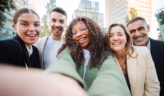 A diverse group of five adults smiling and taking a selfie outdoors in an urban setting with tall buildings in the background.