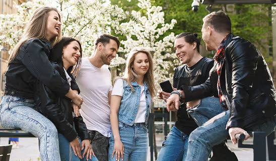 A group of young adults hanging out outdoors, sitting on benches and ledges, laughing and talking together.