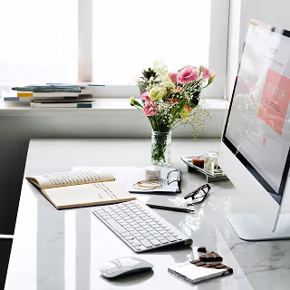 Minimalist marble desk setup with an iMac, wireless keyboard and mouse, open notebook, glasses, pen, and fresh flowers in a vase beside a window.