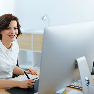 Smiling woman working at a large desktop computer, holding a stylus and appearing focused in a bright, modern office space.