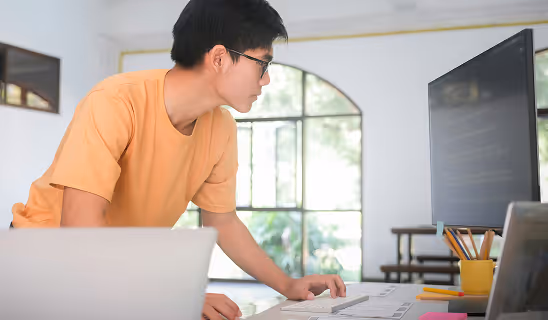 Man in a yellow shirt standing and concentrating on code displayed on a large monitor in a bright, casual workspace.