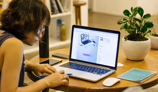 Woman working on a laptop at a small round table, pen in hand, with a potted plant and smartphone beside her.