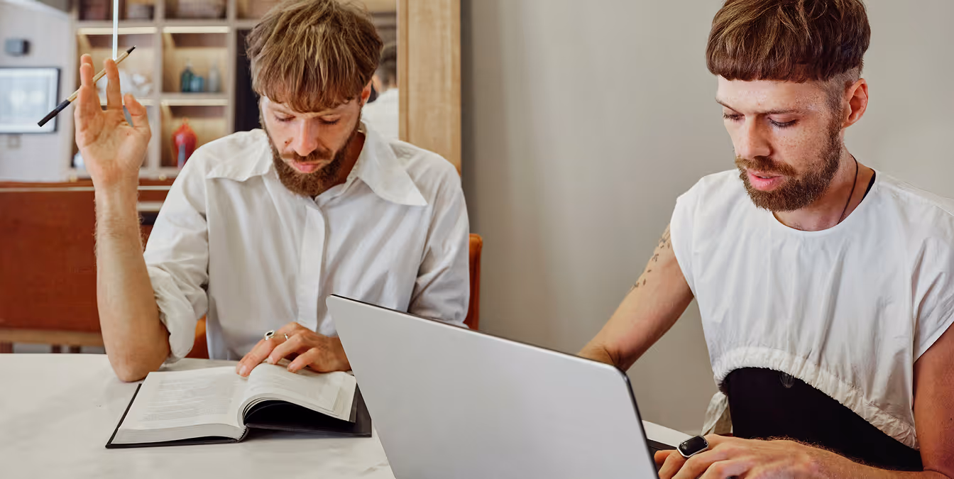 Two men seated at a table, one reading a book and the other working on a laptop, both appearing engaged in a discussion.