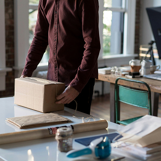 Person in a maroon shirt wrapping a cardboard box on a table with packing materials, preparing a product for shipping.