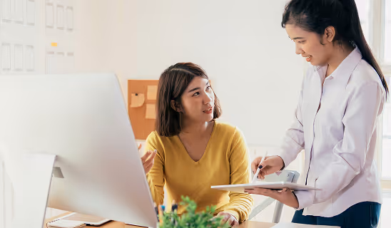 Two women in an office discussing work while standing near a large desktop computer, one holding a clipboard.