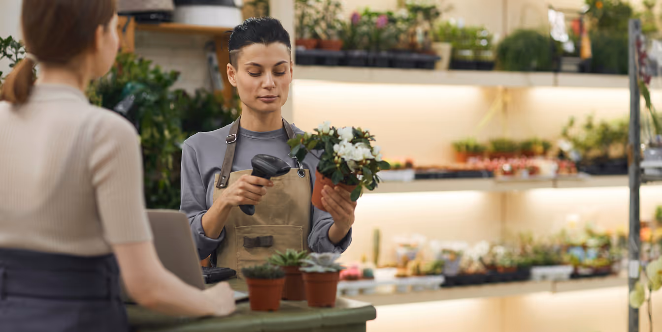 Female florist scanning a potted plant at the checkout counter while speaking with a customer in a well-lit shop filled with greenery.