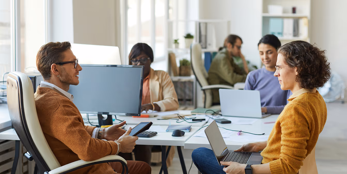 Group of professionals in an open-plan office, two in the foreground conversing with laptops while others work in the background.