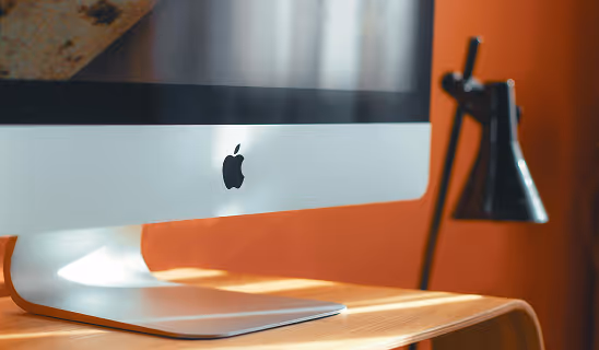 Close-up of an Apple iMac computer on a wooden desk with a black lamp in the background, bathed in warm light.