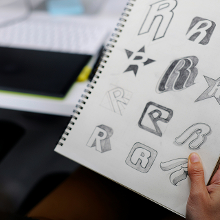 A sketchbook page filled with various hand-drawn letter “R” logo concepts in different styles, including outlined, shaded, and block designs, held by a person in a creative workspace.