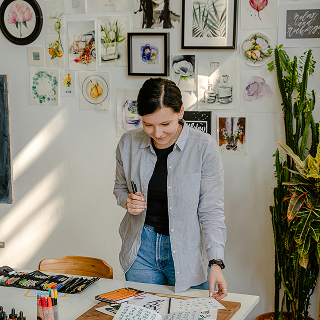 Smiling woman in a casual shirt standing at a desk covered in markers and art supplies, surrounded by a wall display of framed botanical and artistic prints.