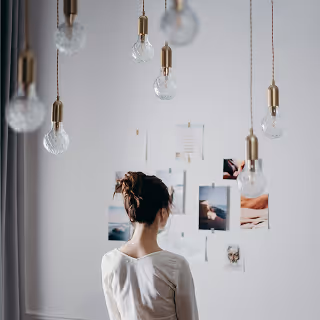 Back view of a woman standing in a softly lit room, looking at a wall collage of photos and artwork, with several hanging light bulbs in the foreground.