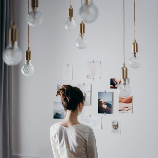 Back view of a woman standing in a softly lit room, looking at a wall collage of photos and artwork, with several hanging light bulbs in the foreground.