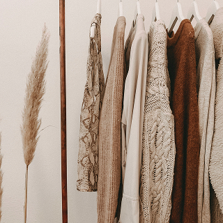 Neutral-toned knit sweaters and patterned tops hanging on a clothing rack beside a dried pampas grass arrangement.