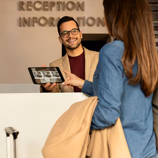 Hotel receptionist smiling and holding a tablet while assisting a guest at the front desk.