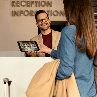 Hotel receptionist smiling and holding a tablet while assisting a guest at the front desk.