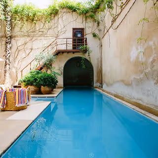 Narrow outdoor swimming pool with stone walls, lush plants, and a striped towel on a chair beside the water.