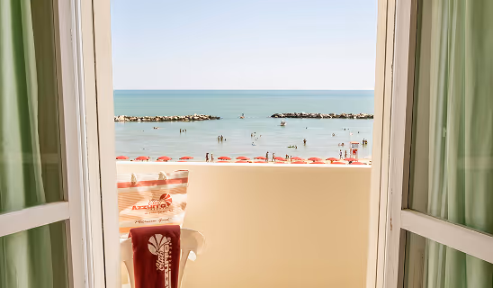 View from a hotel room balcony overlooking a beach and ocean, with red umbrellas and people swimming.