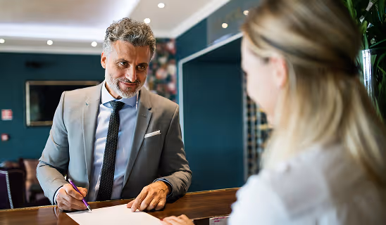 Hotel receptionist in a suit smiling as a guest signs paperwork at the front desk.