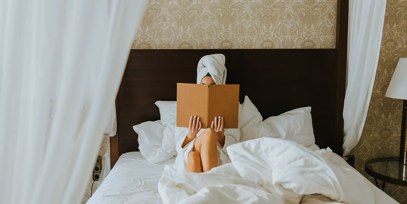 Person relaxing in a hotel bed with a towel wrapped around their head, reading a menu or book, surrounded by white sheets and draped curtains.