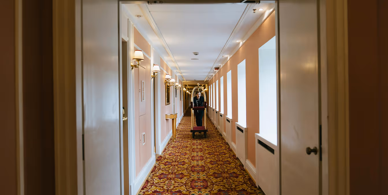 Hotel staff member pushing a luggage cart down a long hallway with patterned carpet and pink walls lined with windows. Ask ChatGPT