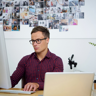 A man wearing glasses and a red checkered shirt works intently at a large desktop computer, with a laptop and a wall covered in photos behind him.