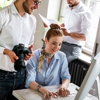 A smiling woman types at a desktop computer while a man beside her holds a camera, and another colleague reads papers in the background.