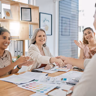 Two women shake hands across a table covered in documents and color swatches, while colleagues smile and applaud in a bright office.