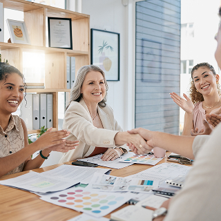 Two women shake hands across a table covered in documents and color swatches, while colleagues smile and applaud in a bright office.