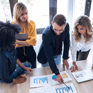 A group of professionals gather around a table, examining printed graphs and charts during a collaborative strategy discussion.