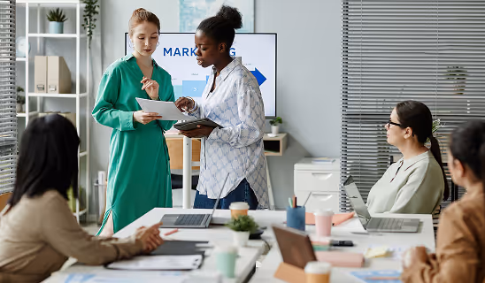 Two women stand in front of a screen displaying the word “Marketing,” reviewing notes while team members listen at a meeting table.