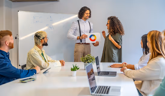 A man holding a brand logo printout presents to colleagues in a meeting room with laptops and a whiteboard.