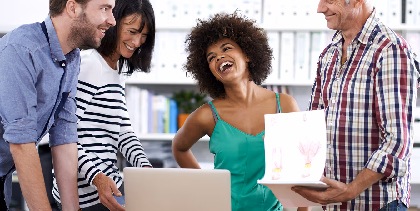 A group of four colleagues stand around a laptop in a bright office, smiling and laughing while reviewing printed designs.