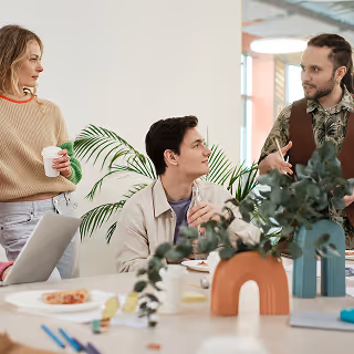 Casual office meeting with three colleagues talking around a desk, with plants and laptops in the background.