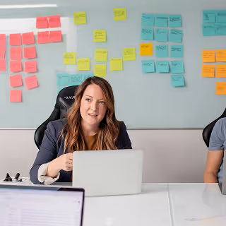 Woman speaking in a meeting room with a laptop, while a whiteboard covered in colorful sticky notes is visible behind her.