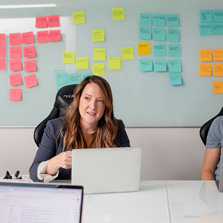 Woman speaking in a meeting room with a laptop, while a whiteboard covered in colorful sticky notes is visible behind her.