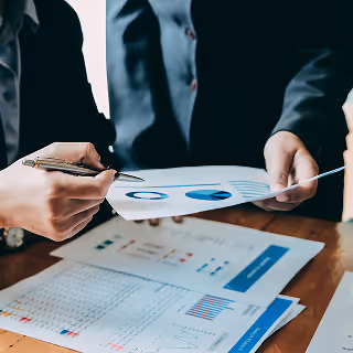 Two people in suits reviewing printed charts and graphs on a table, discussing marketing or business data.