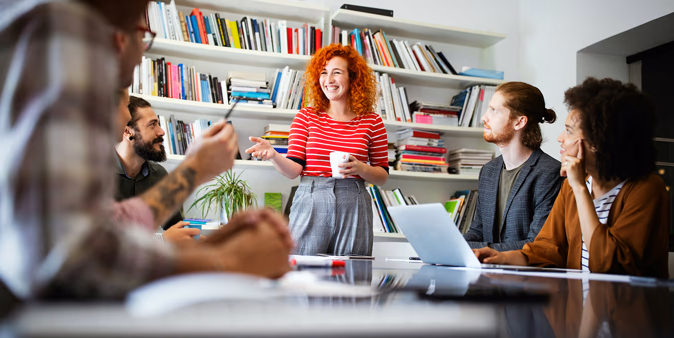 Woman with curly red hair leading a lively team discussion in an office with bookshelves, as colleagues listen attentively.