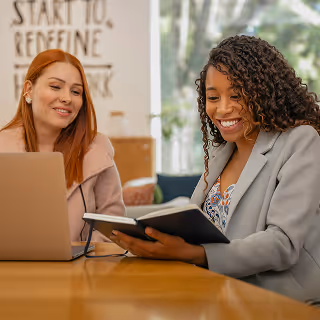 Two women sitting at a desk, smiling while reviewing notes together, with a laptop in front of them.