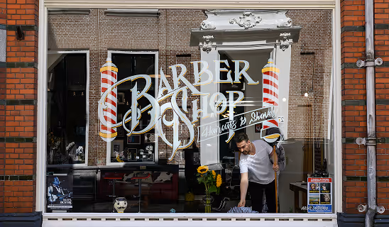 View through a barbershop window showing a barber preparing a chair inside the shop.