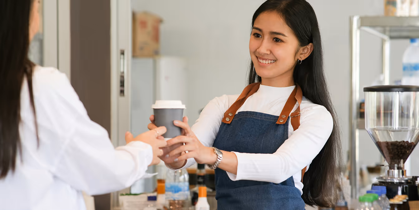 Smiling barista in an apron handing a takeaway coffee cup to a customer inside a café.