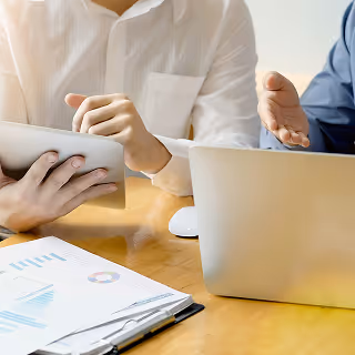 Two people using a tablet and laptop while discussing graphs and charts during a meeting.