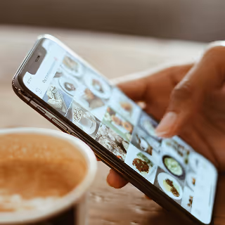 Close-up of a hand scrolling through a smartphone gallery of food images, with a cup of coffee in the background.