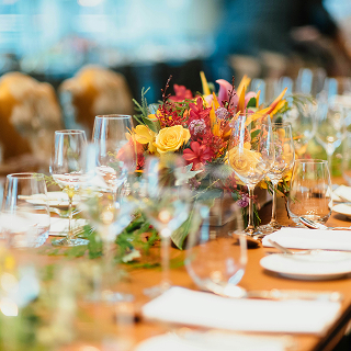 Long dining table set with wine glasses, plates, and a vibrant floral centerpiece in warm lighting.