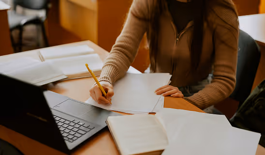 Person writing on paper at a desk with open books and a laptop, focusing on study or research work.