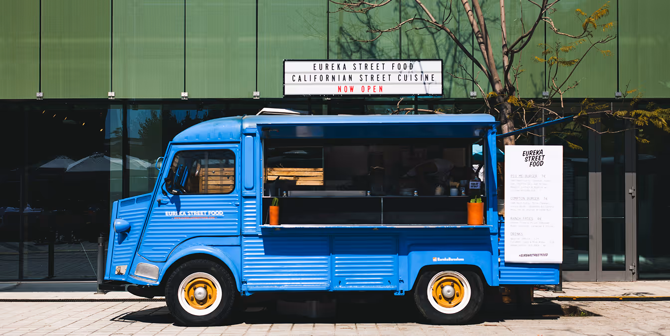Bright blue food truck parked outside a modern building, with a large menu board and a sign reading “Now Open.”