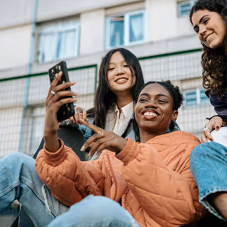 Young women sitting together, smiling and posing for a selfie, with a building in the background.